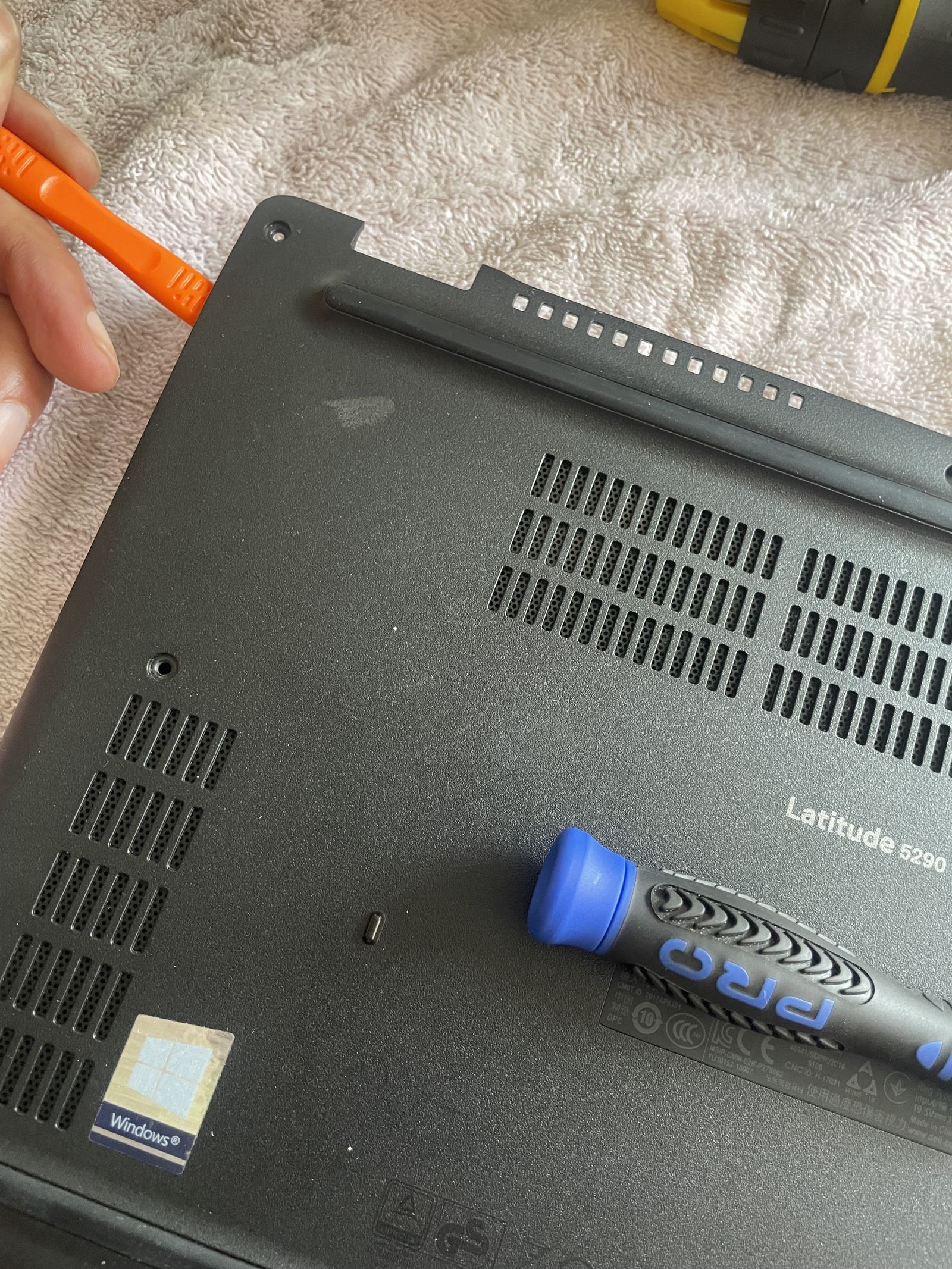 A hand working an orange plastic pry tool along the seam of the laptop’s base, next to the rear vents.