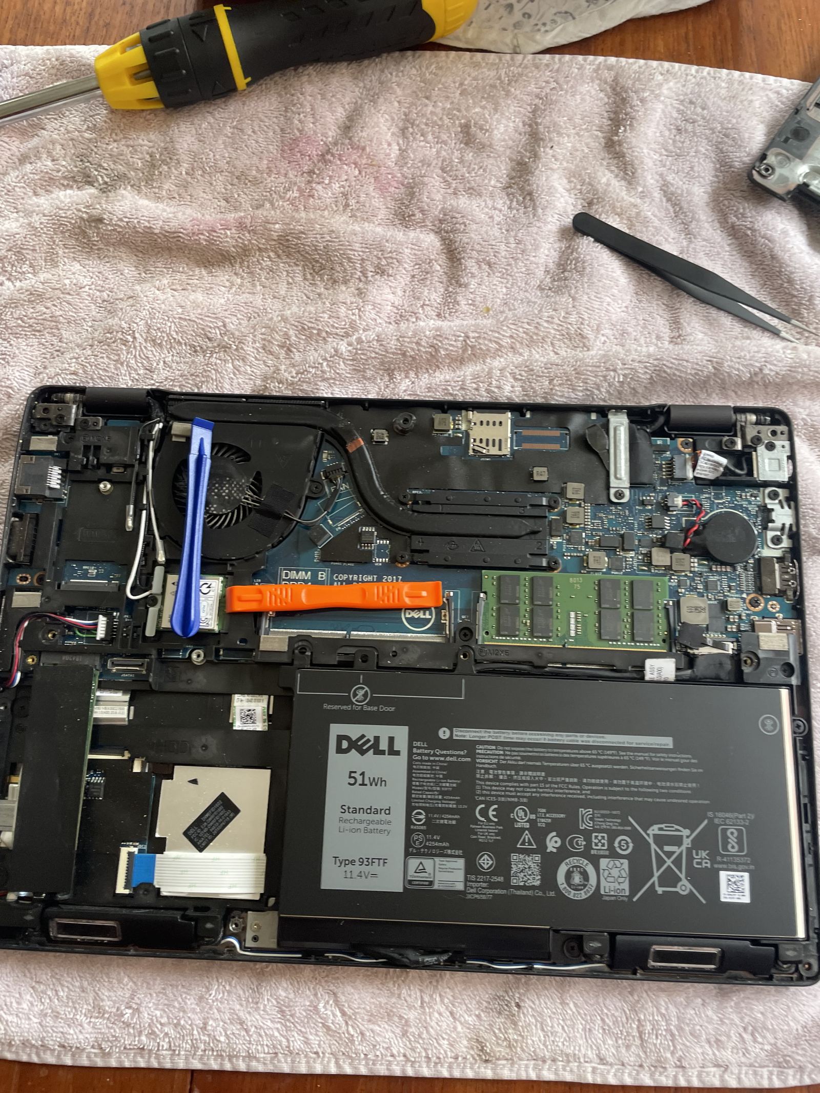 The laptop with its base off and the new 51 Wh battery fitted, a blue and an orange plastic pry tool resting on the board, tweezers on the towel above.
