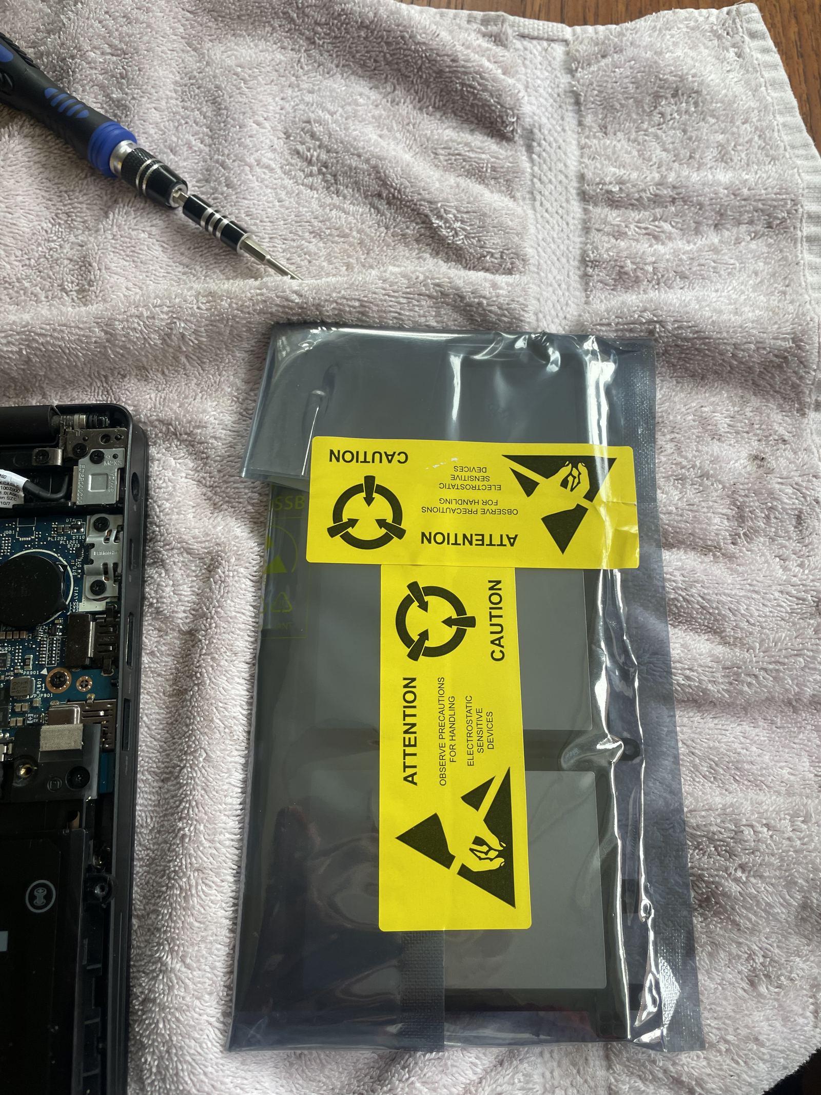 The replacement battery still sealed in its silver anti-static bag with yellow caution labels, lying on the towel beside the open laptop.