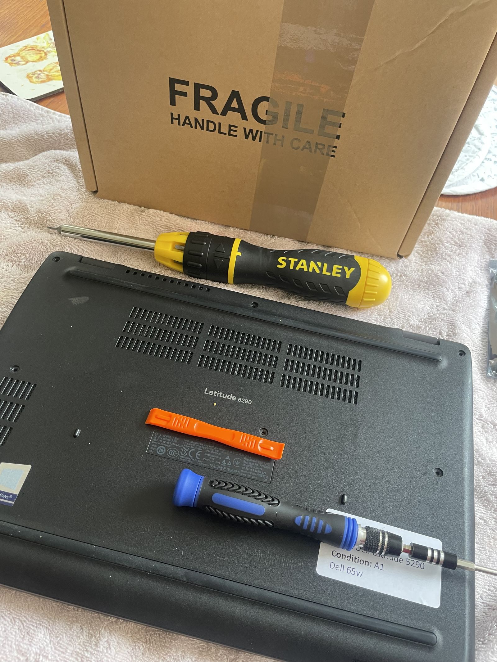 The laptop upside down on a towel, with a Stanley ratchet driver, a precision driver, an orange plastic pry tool and the boxed replacement part behind it.