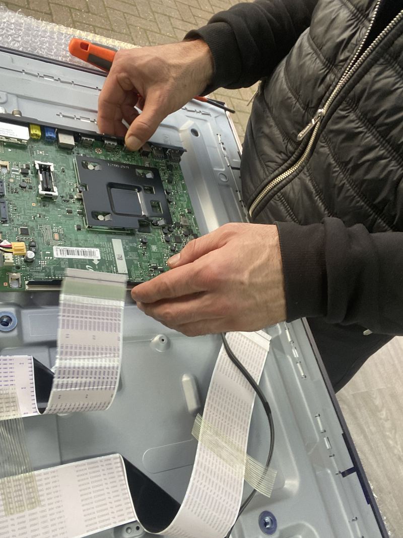 Two hands working on the green mainboard of a television lying face down with bubble wrap beneath one edge, ribbon cables disconnected and hanging loose, a screwdriver held in one hand.