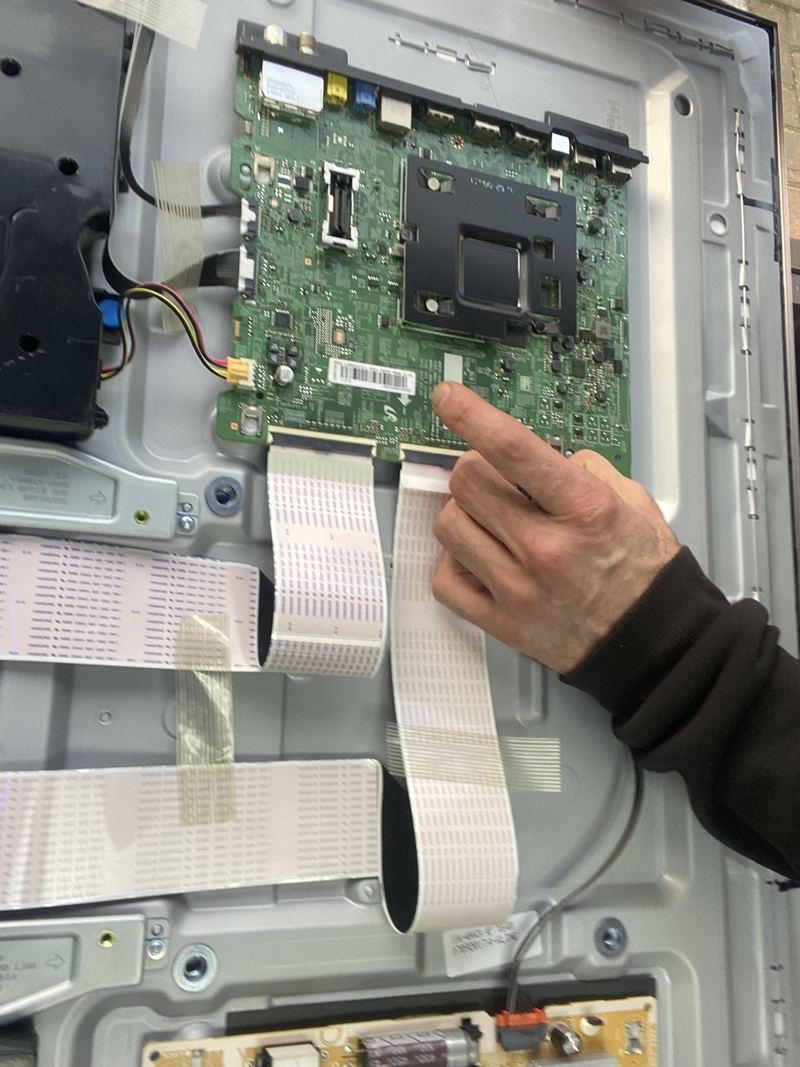 A technician's hand pointing at the mainboard inside an opened television, with the panel's ribbon cables fanned out below it.