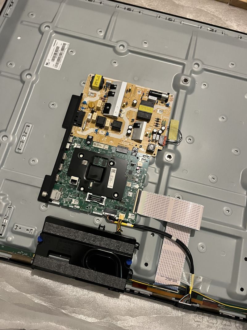 The rear chassis of a television with its back cover removed, the cream power board and green mainboard mounted side by side, a black speaker enclosure below and ribbon cables running to the panel.
